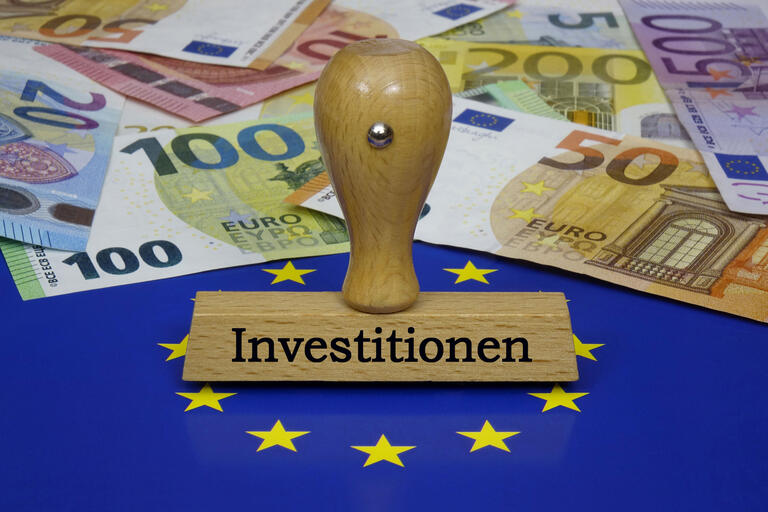 Auf einer Europafahne liegen Geldscheine und shttps://prod-cms1.boeckler.de/cms/AssetManager/#teht ein Stempel mit der Aufschrift Investitionen. 
