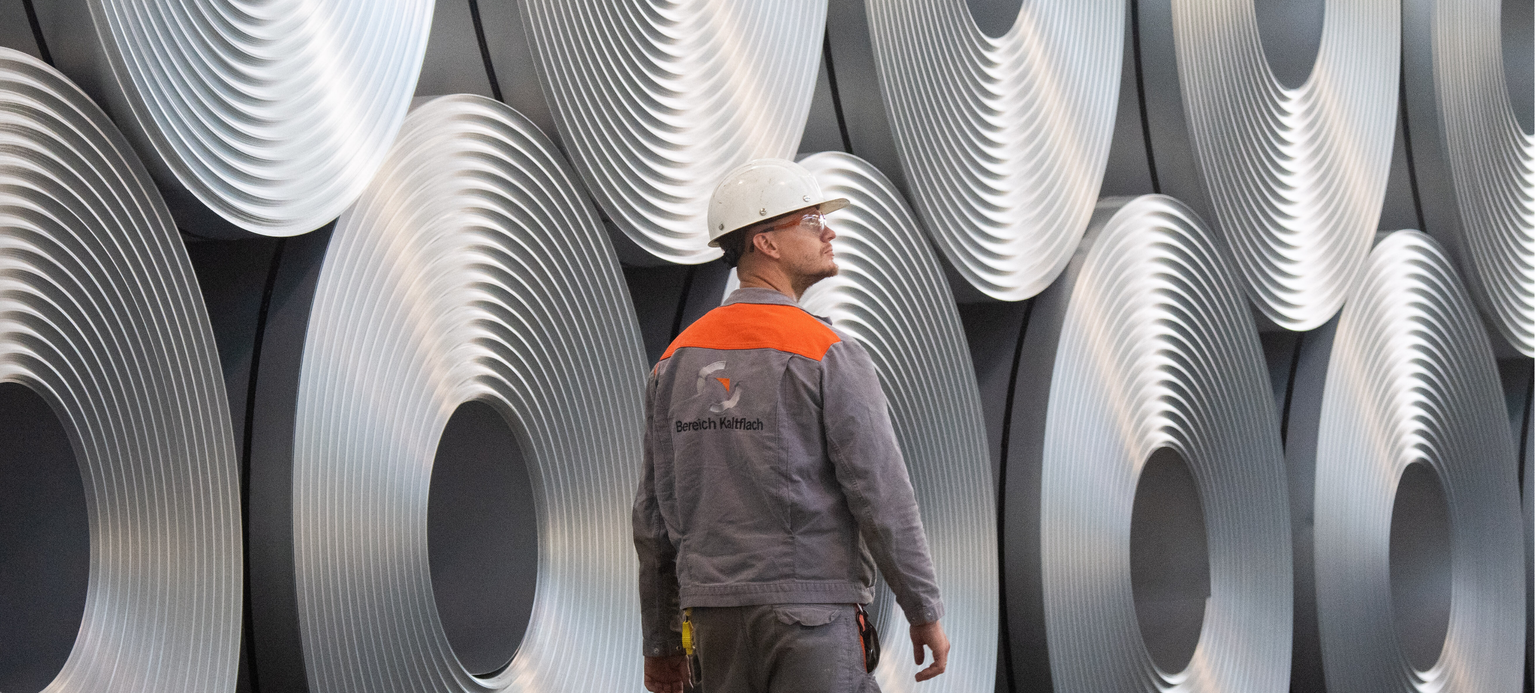 Eine Mitarbeiter geht bei der Salzgitter AG an aufgewickeltem Stahl (Coils) vorbei. Der Stahlhersteller Salzgitter AG will den CO2-Ausstoß erheblich senken. Als Teil der Klimastrategie des Unternehmens soll Kohle bei der Erzeugung von Eisen schrittweise durch Wasserstoff und Strom aus erneuerbaren Quellen ersetzt werden. 