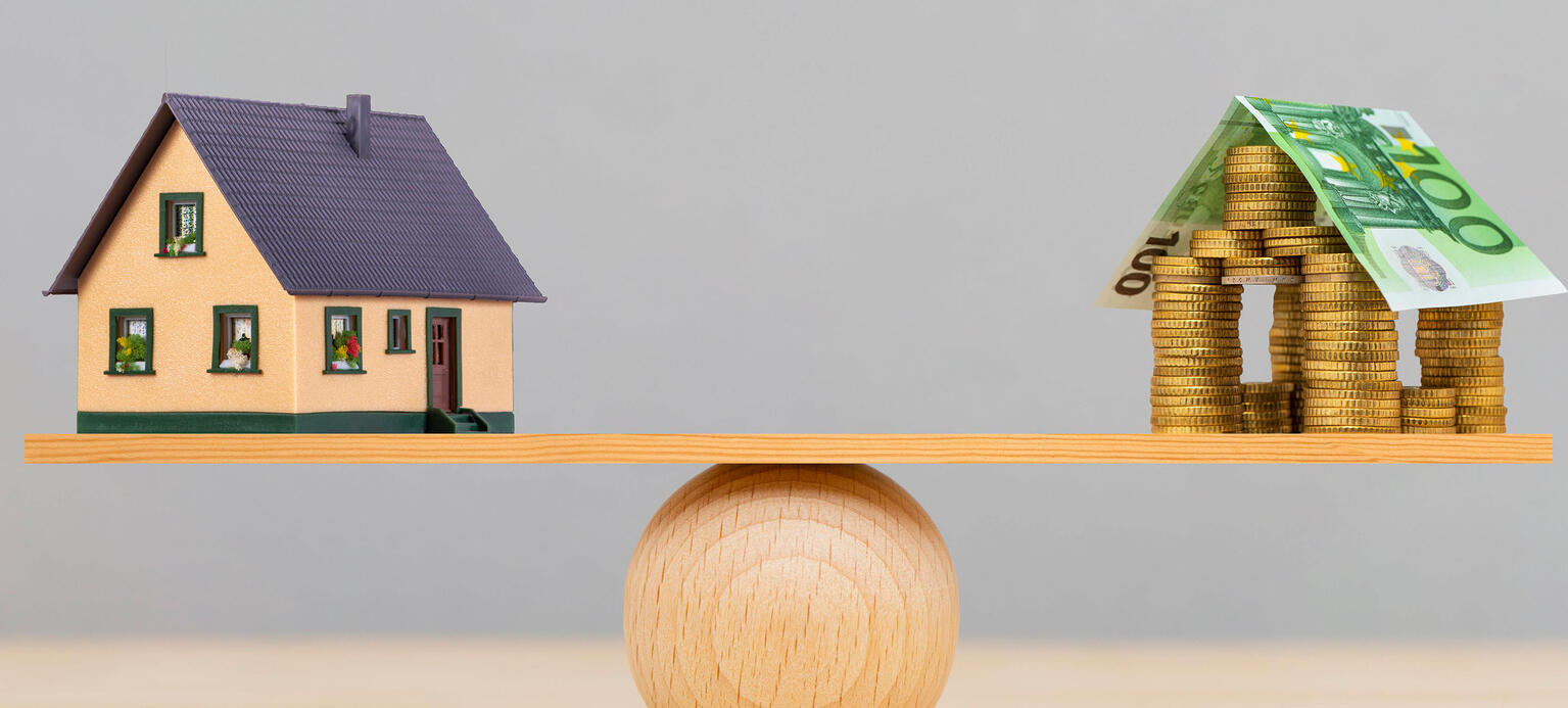Waage aus Holz mit einem Modell Haus auf der einen Seite und einem Haus aus Geldscheinen auf der Anderen. Sie halten sich die Waage