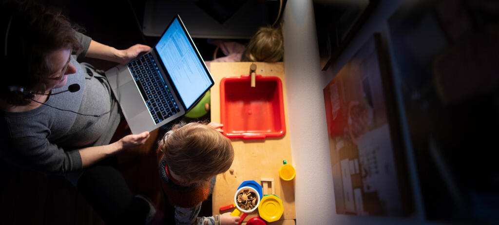 Eine Frau arbeitet am Laptop. Neben ihr steht ein Kleinkind an einer Kinderküche.