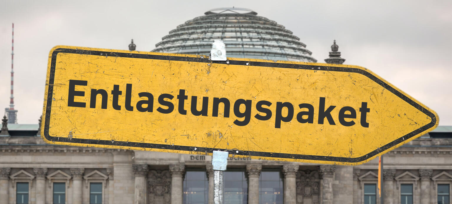 Ein gelbes, zerkratztes Hinweisschild mit der Aufschrift 'Entlastungspaket' vor dem Berliner Reichstagsgebäude