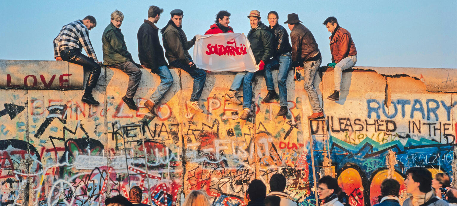 Nach Öffnung der DDR-Grenzen zum Westen (9.11.1989): Eine Gruppe von Menschen sitzt auf der Berliner Mauer und hält ein Plakat mit der Aufschrift „Solidarnosc“ hoch.