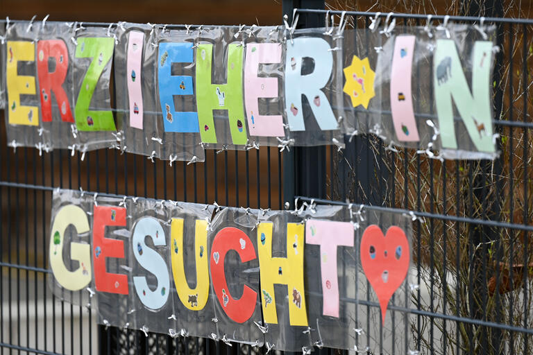Mit bunten Buchstaben sind die Worte „Erzieher*in gesucht“ geformt, die an einem Zaun einer Kindertagesstätte befestigt sind