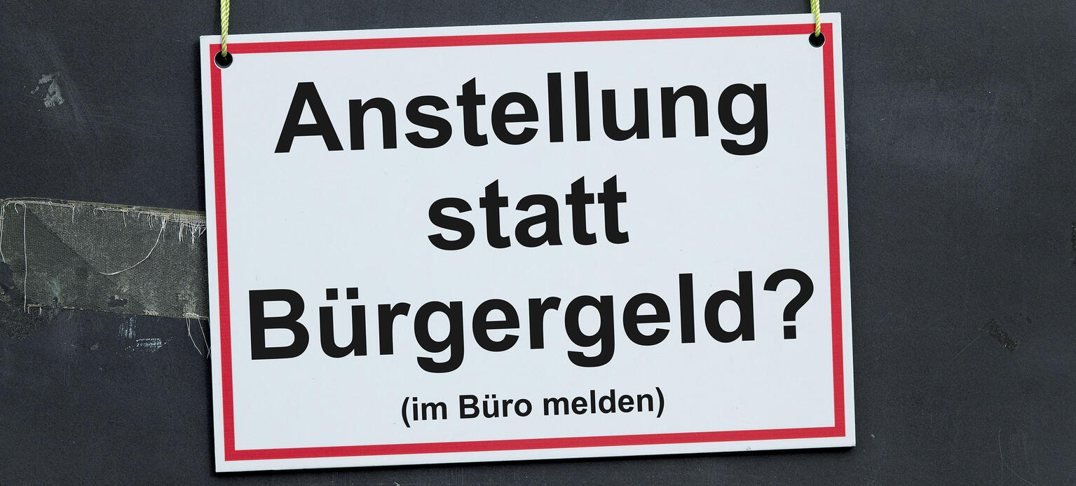 Ein weisses Schild mit rotem Rand und der Aufschrift 'Anstellung statt Buergergeld? (im Büro melden)' haengt schief an gelben Schnueren vor einer Tafel mit Resten von Klebeband.