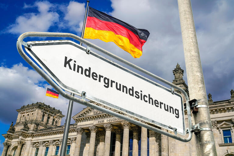 Ein weisses Hinweisschild an einem Laternenmast mit abgeblaetterter Farbe am Rand und der Aufschrift 'Kindergrundsicherung' als Wegweiser nach links vor dem Berliner Reichstagsgebaeude mit wehenden Deutschlandflaggen