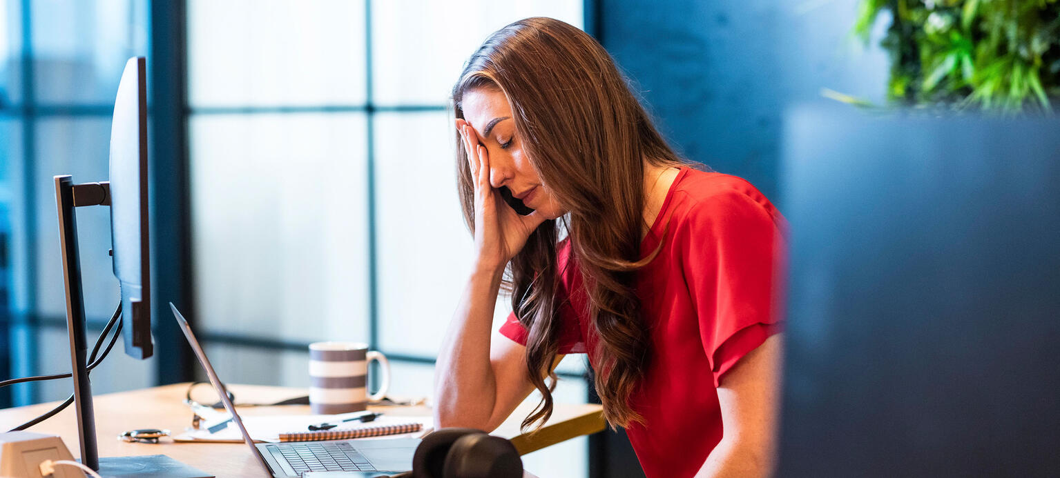 Eine Frau sitzt an einem Tisch und hält die Hand vor ihr Gesicht. Auf dem Tisch stehen diverses Büroequipment, wie ein Laptop, ein Bildschirm und ein Headset. Sie sieht verzweifelt aus.