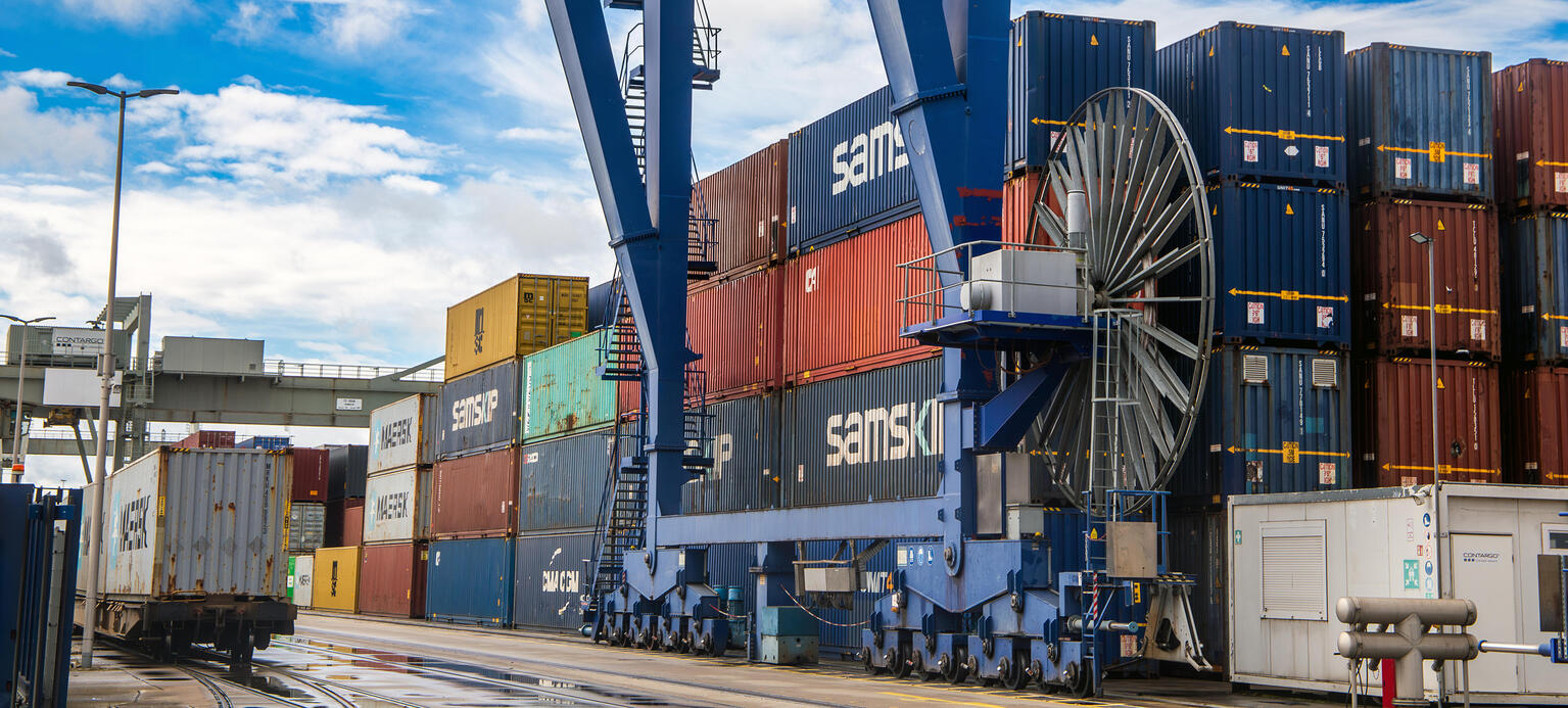 Ein Containerhafen in dem ein großer Kran die einzelnen Container unter blauem, leicht wolkigen Himmel bewegt.