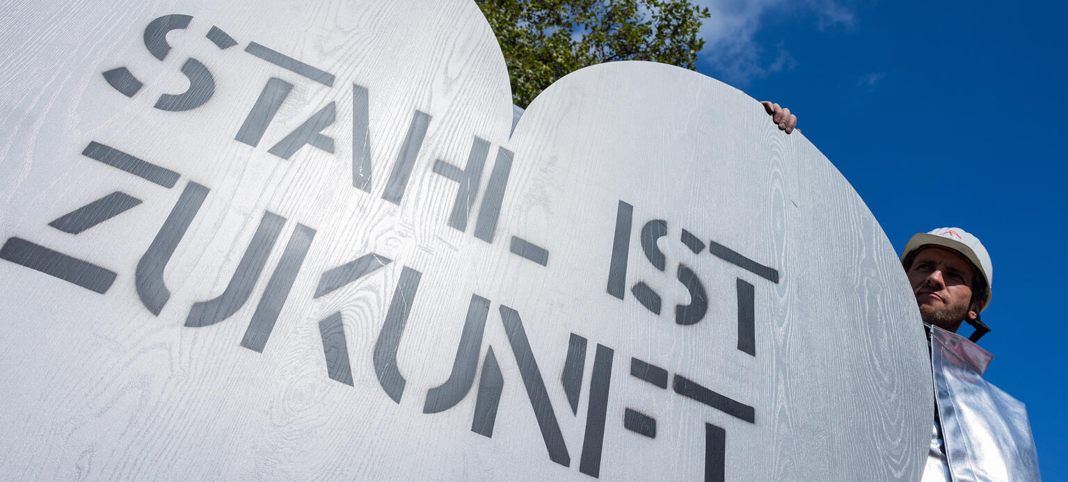 Stahlarbeiter demonstrieren einem überdimensionalen grauen Herz mit der Aufschrift 'Stahl ist Zukunft'.