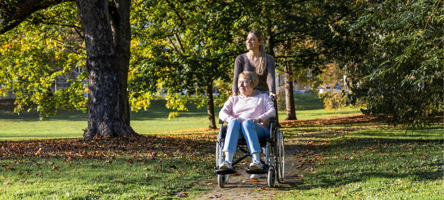 Eine junge Krankenschwester fährt eine ältere Dame im Rollstuhl durch den Park.