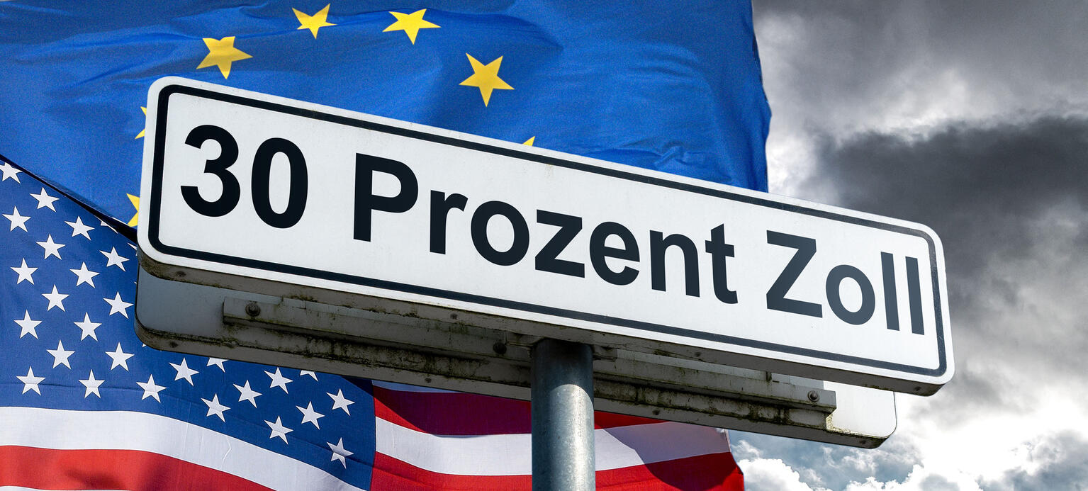 Symbolbild: ein weisses Schild mit schwarzer Umrandung und schwarzer Aufschrift '30 Prozent Zoll' an einem Schildermast von schraeg unten vor der im Wind wehenden Nationalflagge der Vereinigten Staaten von Amerika und der Flagge der Europaeischen Union an einem grau bewoelkten Himmel. 