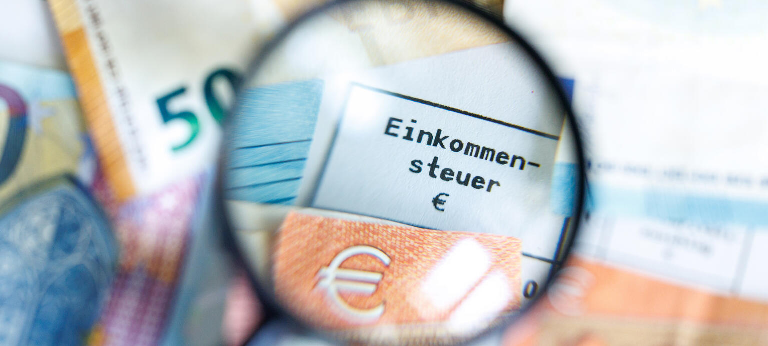 Der Begriff Einkommensteuer ist durch eine Lupe auf einem Steuerbescheid vom Finanzamt zu sehen, auf dem verschiedene Banknoten liegen.