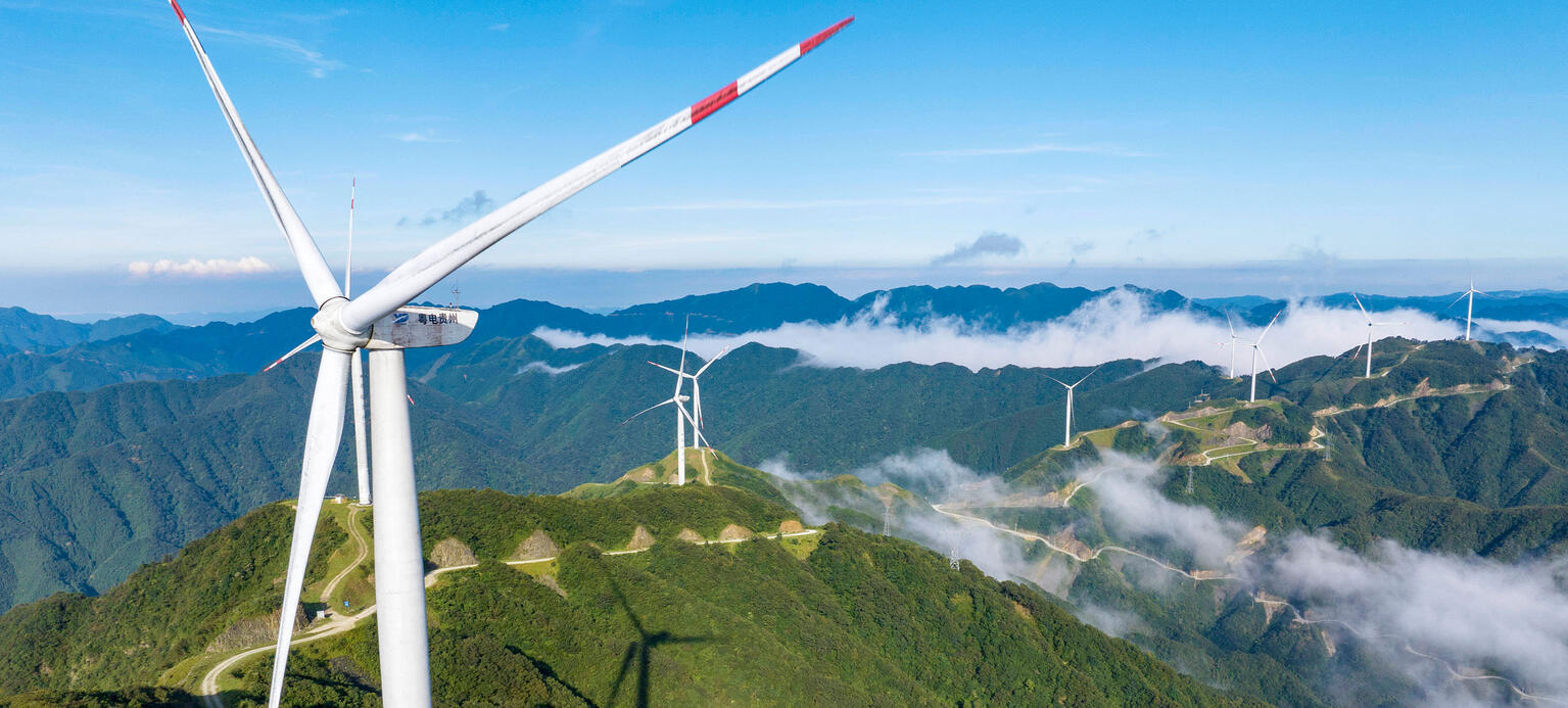 Symbolbild: Windräder stehen auf begrünten Bergen. Es ist ein blauer Himmel mit vereinzelten Wolken im Hintergrund zu sehen.