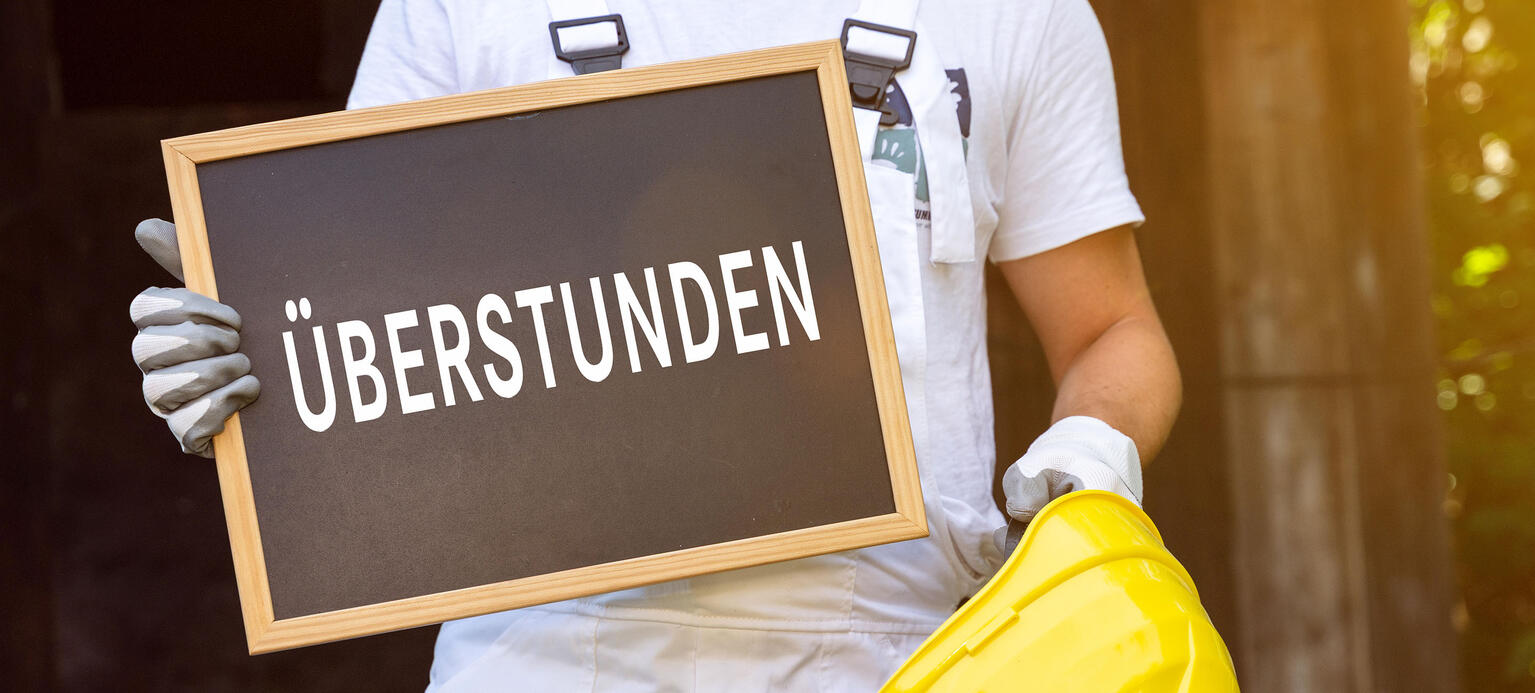 Fotomontage, Ein Arbeiter im weißen Overall hält ein Schild mit der Aufschrift Überstunden sowie einen gelben Bauhelm. Das Motiv symbolisiert zusätzliche Arbeitszeiten im Handwerk und auf Baustellen.