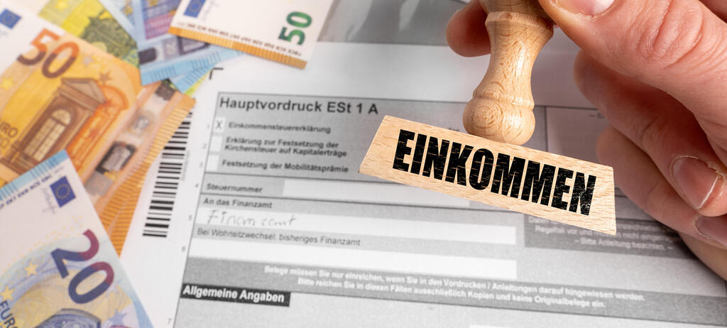 Fotomontage: Ein Stempel mit der Aufschrift Einkommen wird über einer Einkommensteuererklärung gehalten. Euro-Banknoten daneben symbolisieren Einkommen und steuerpflichtige Einkünfte im Zusammenhang mit der Einkommensteuer.