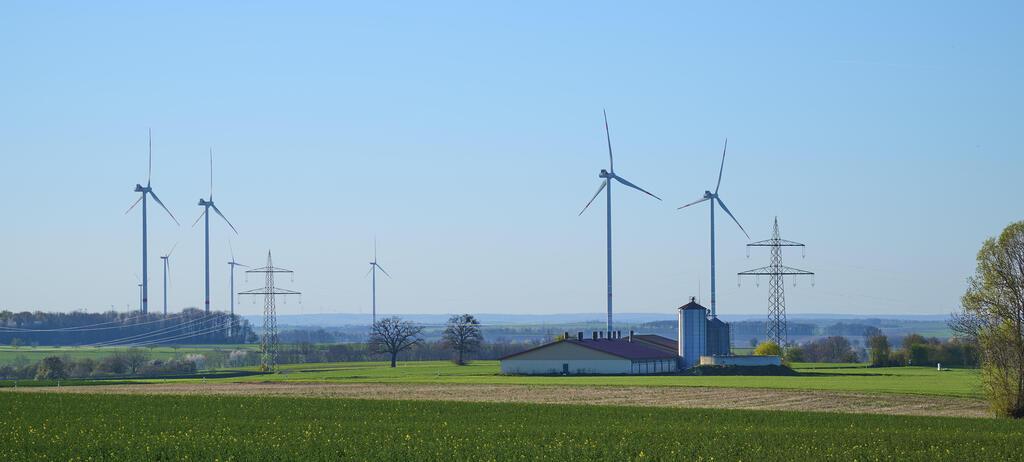 Windräder und Stromleitungen auf einer grünen Wiese mit einem Bauernhof unter klarem Himmel.