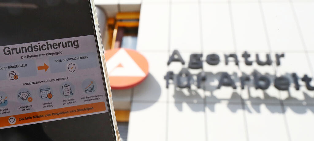 Ein Smartphone zeigt eine Grafik zur neuen Grundsicherung als Nachfolgeregelung des Bürgergeldes. Im Hintergrund ist das Gebäude der Bundesagentur für Arbeit zu sehen. 