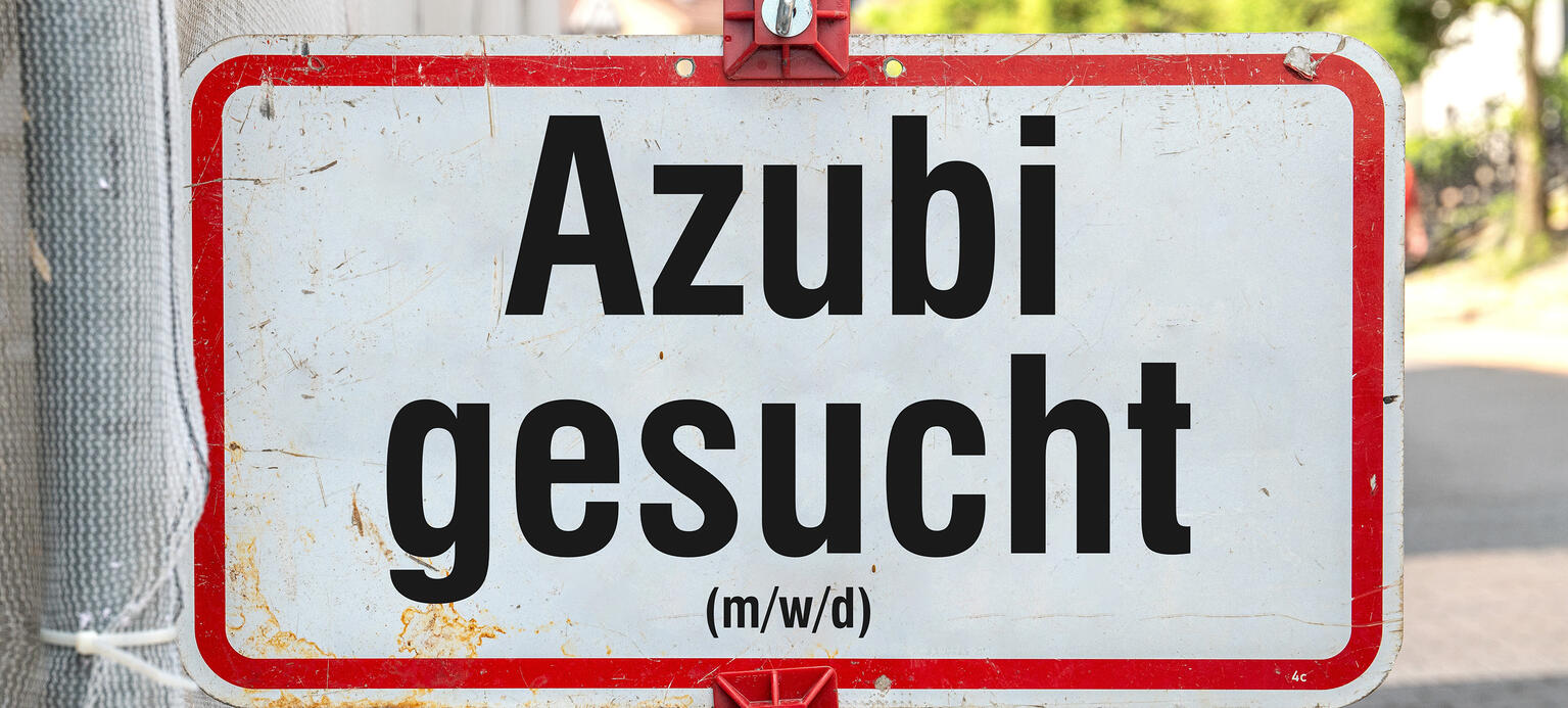 Symbolbil: ein weisses Schild mit roter Umrandung und schwarzer Aufschrift 'Azubi gesucht (m/w/d)' an einem Schildermast mit roten Haltern vor einem Haus mit Baustelle. 