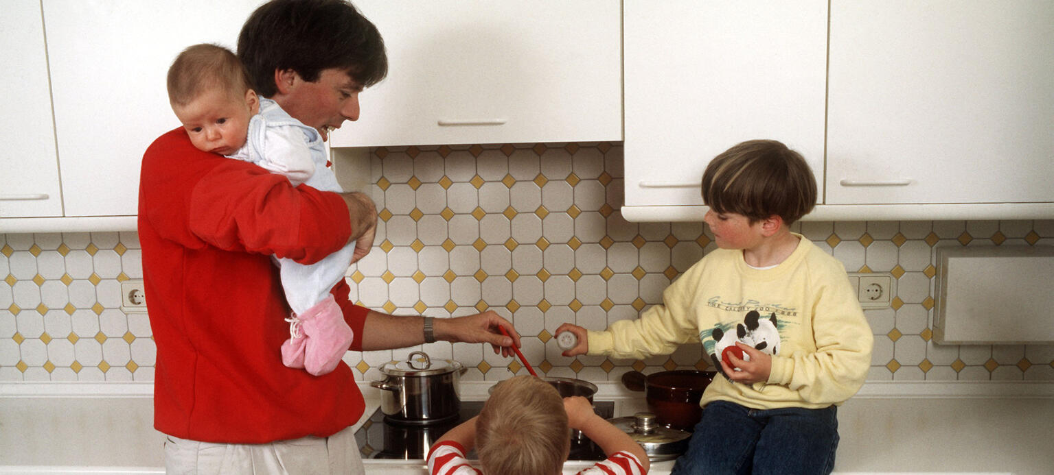 Ein Vater, der einen Säugling auf dem Arm trägt, bekommt von seinen zwei größeren Kindern Hilfe beim Kochen, aufgenommen 1988. 