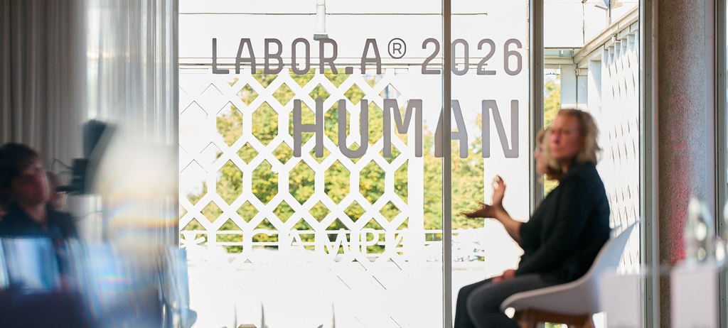 Eine Glasscheibe mit dem Schriftzug 'Labora 2026 HUMAN', davor sind unscharf zwei Personen zu erkennen, die gestikulieren