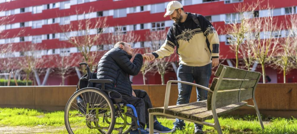 Ein Mann in einem Rollstuhl und ein Mann, der ihn unterstützt stehen vor einem großen Gebäude auf einer Wiese. Der Mann im Rollstuhl hält die Hand des Betreuers